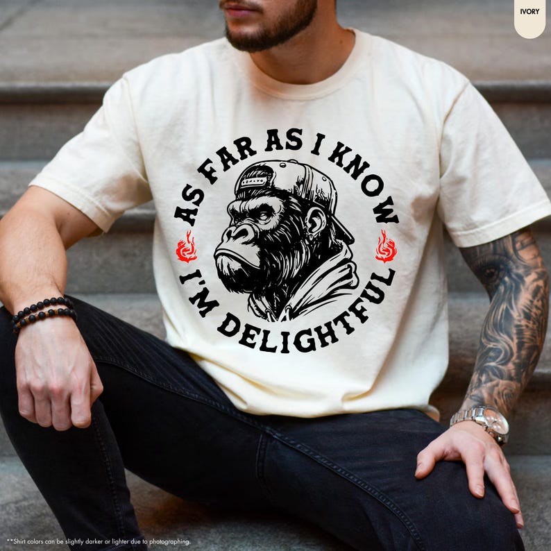 May include: Ivory-colored t-shirt featuring a black and white graphic of a gorilla wearing a cap. The text around the gorilla reads "AS FAR AS I KNOW I'M DELIGHTFUL" in a circular design. The shirt is a casual wear item.