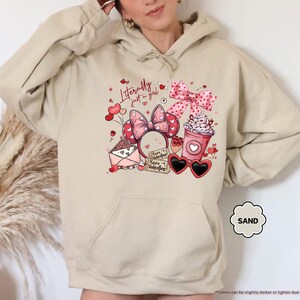 May include: Sand-coloured hoodie featuring a Valentine's Day design. The graphic includes a pink bow, heart-shaped sunglasses, a coffee cup, and the text "Literally just a girl." The hoodie has a drawstring hood and a kangaroo pocket.
