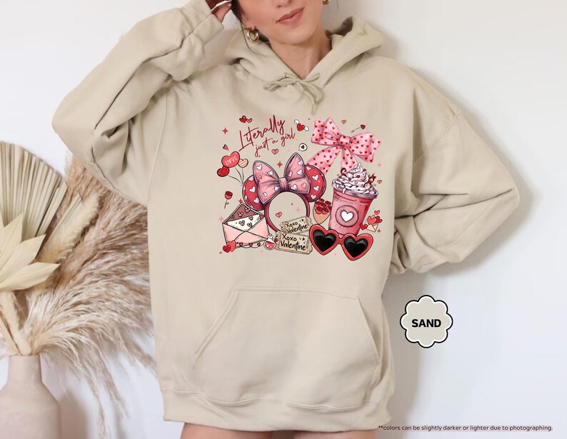 May include: Sand-coloured hoodie featuring a Valentine's Day design. The graphic includes a pink bow, heart-shaped sunglasses, a coffee cup, and the text "Literally just a girl." The hoodie has a drawstring hood and a kangaroo pocket.