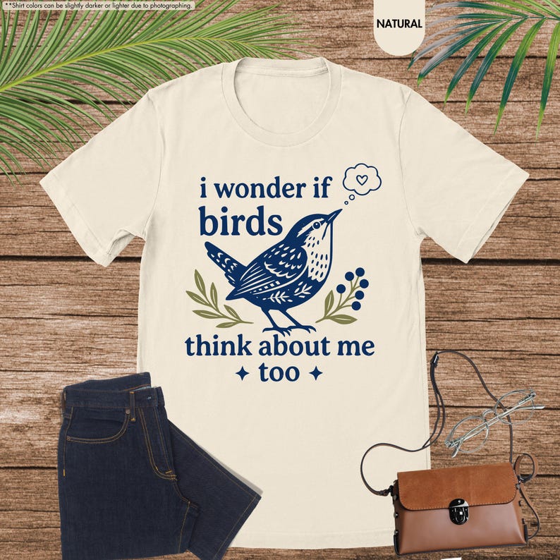 May include: A natural-colored t-shirt featuring a dark blue graphic of a bird perched on a branch with berries. The text reads, "i wonder if birds think about me too" with a small thought bubble containing a heart icon above the bird.