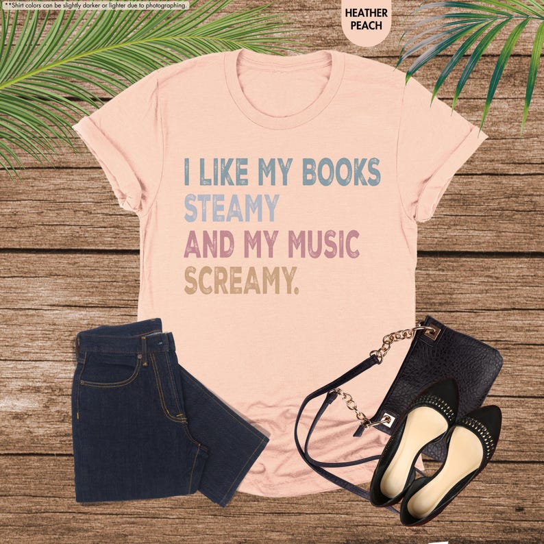 May include: A heather peach t-shirt with the text "I LIKE MY BOOKS STEAMY AND MY MUSIC SCREAMY." The shirt is paired with dark wash jeans, a black purse, and black flats with gold studs. The items are arranged on a wooden surface.