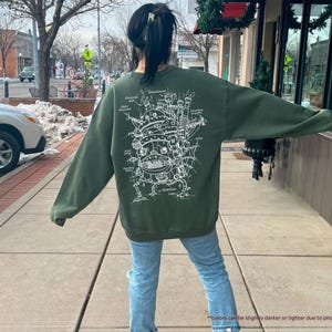 May include: A green sweatshirt with a white graphic print of a flying machine with intricate details. The graphic is a detailed line drawing of a flying machine with a large central structure and multiple smaller structures attached to it. The sweatshirt is a casual style with a crew neck and long sleeves.