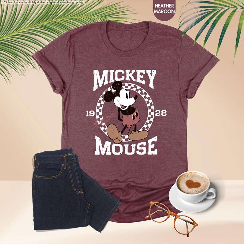May include: Heather maroon t-shirt featuring a graphic of Mickey Mouse with the text "MICKEY MOUSE 1928". The shirt is paired with dark wash denim jeans, a cup of coffee with a heart design, and eyeglasses.