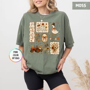 May include: A moss green t-shirt featuring a graphic print with the text hello fall. The design includes a tractor, a duck wearing a hat, floral arrangements, pumpkins, and a plaid bow, all arranged in rectangular and oval frames on the front.