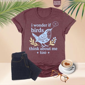May include: A heather maroon short-sleeve t-shirt featuring a light blue graphic of a bird and the text "i wonder if birds think about me too" with a small heart in a thought bubble, accompanied by dark blue jeans, glasses, and a coffee cup.