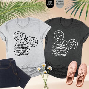 May include: Two t-shirts with a Mickey Mouse ear design and the text "I'm done adulting I'm going to Disney". The shirt on the left is light grey heather and the shirt on the right is dark grey heather.