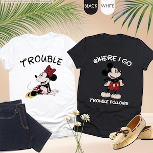 May include: Two t-shirts, one white and one black, featuring Disney characters. The white shirt has Minnie Mouse with the word "TROUBLE". The black shirt has Mickey Mouse with the text "WHERE I GO TROUBLE FOLLOWS". Also shown are jeans and shoes.