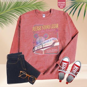 May include: Heather scarlet red sweatshirt with a graphic that reads "PLEASE STAND CLEAR of the Doors" featuring a monorail and fireworks. The image also includes a pair of dark wash jeans, red sneakers, and eyeglasses.