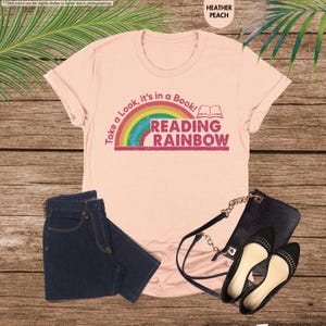 May include: A peach-colored t-shirt with the text "Take a Look, it's in a Book! READING RAINBOW" and a rainbow graphic. The image also shows a pair of dark blue jeans, a black purse, and black flats with studs.