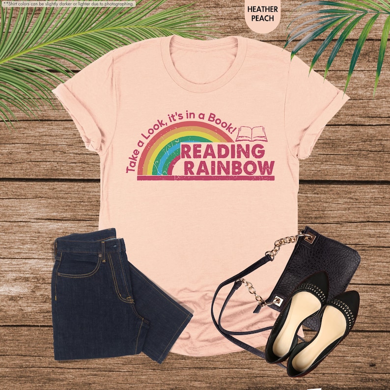 May include: A peach-colored t-shirt with the text "Take a Look, it's in a Book! READING RAINBOW" and a rainbow graphic. The image also shows a pair of dark blue jeans, a black purse, and black flats with studs.