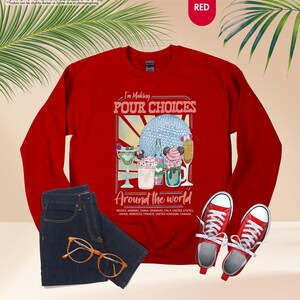 Pu&ograve; includere: Una felpa rossa con la scritta "I'm Making POUR CHOICES Around the world" e una grafica di bevande. L'immagine include jeans blu, occhiali con montatura dorata e scarpe da ginnastica rosse.