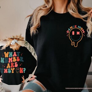 以下が含まれることがあります： カラフルなスマイリーフェイスのグラフィックと「Dance Mom」の文字が入った黒いスウェットシャツ。スウェットシャツの背面には、「What number are they on?」という文字がカラフルな文字で書かれています。