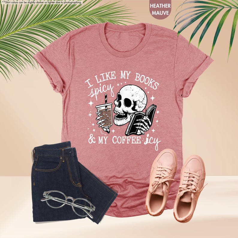 May include: A heather mauve t-shirt with a graphic of a skull holding a book and a cup of coffee. The text on the shirt reads "I like my books spicy & my coffee icy".