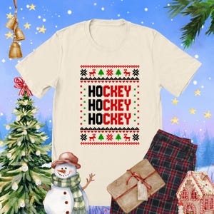 May include: A white t-shirt with a red, green, and black Christmas sweater design. The design features the word "HOCKEY" repeated three times.