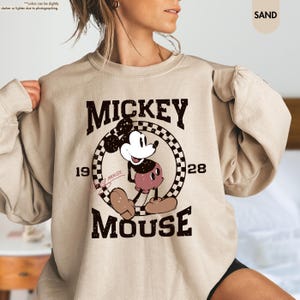 May include: Sand-colored sweatshirt featuring a vintage Mickey Mouse graphic. The design includes the words "MICKEY MOUSE" and the year "1928" in a distressed font. The graphic is centered on the chest, with Mickey Mouse in a classic pose.