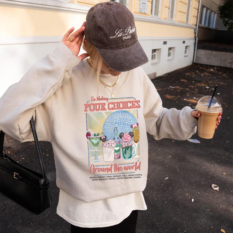 Pu&ograve; includere: Una felpa beige con la scritta "I'm Making POUR CHOICES Around the world" e una grafica di bevande e una sfera blu. Si indossa un berretto da baseball marrone con "La Paris". Sono visibili anche una borsa nera e una bevanda al caff&egrave;.