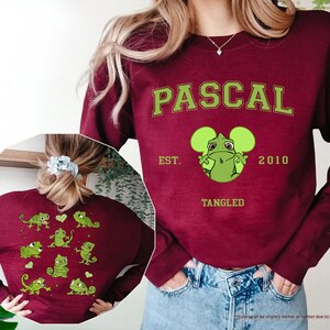 Op de afbeelding: Bordeaux sweatshirt met het woord "PASCAL" in groene blokletters, een groene cartoonkameleon en de tekst "EST. 2010 TANGLED." De achterkant van de sweatshirt heeft groene kameleongraphics. De sweatshirt wordt gedragen met een blauwe spijkerbroek.