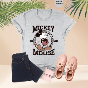May include: A light gray t-shirt featuring a vintage Mickey Mouse graphic, paired with dark blue jeans, pink sneakers, and a small pink wallet. The shirt has the text "MICKEY MOUSE 1928".