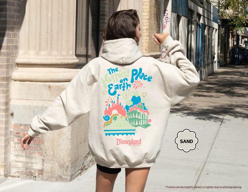 Pu&ograve; includere: Una felpa con cappuccio color crema con una stampa grafica di un castello e fuochi d'artificio. Il testo "The Happiest Place on Earth" e "Disneyland" sono stampati sulla felpa.