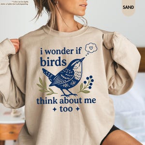 May include: A sand-colored crewneck sweatshirt featuring a dark blue graphic of a bird with a thought bubble containing a heart. The text reads, "i wonder if birds think about me too." The design includes small green leaf and berry accents.