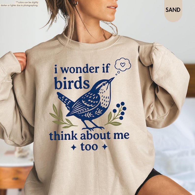 May include: A sand-colored crewneck sweatshirt featuring a dark blue graphic of a bird with a thought bubble containing a heart. The text reads, "i wonder if birds think about me too." The design includes small green leaf and berry accents.