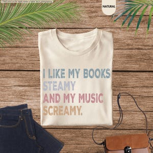 May include: Cream-colored t-shirt with the text "I LIKE MY BOOKS STEAMY AND MY MUSIC SCREAMY" in blue, light blue, pink, and tan. The shirt is laid on a wooden surface with a pair of blue jeans and a brown purse.