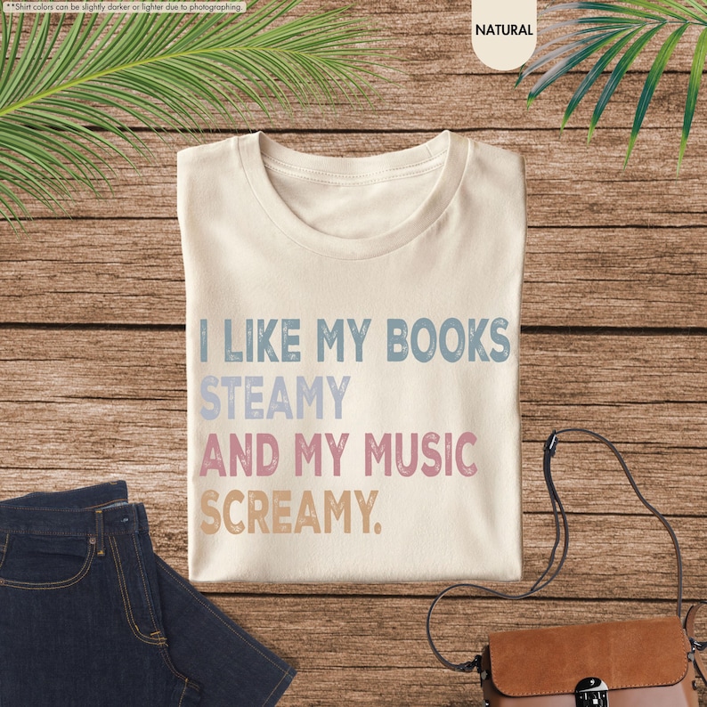 May include: Cream-colored t-shirt with the text "I LIKE MY BOOKS STEAMY AND MY MUSIC SCREAMY" in blue, light blue, pink, and tan. The shirt is laid on a wooden surface with a pair of blue jeans and a brown purse.