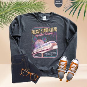 May include: Dark heather gray sweatshirt with a graphic that reads "PLEASE STAND CLEAR of the Doors" featuring a monorail and fireworks. The sweatshirt is paired with dark wash jeans and orange sneakers.