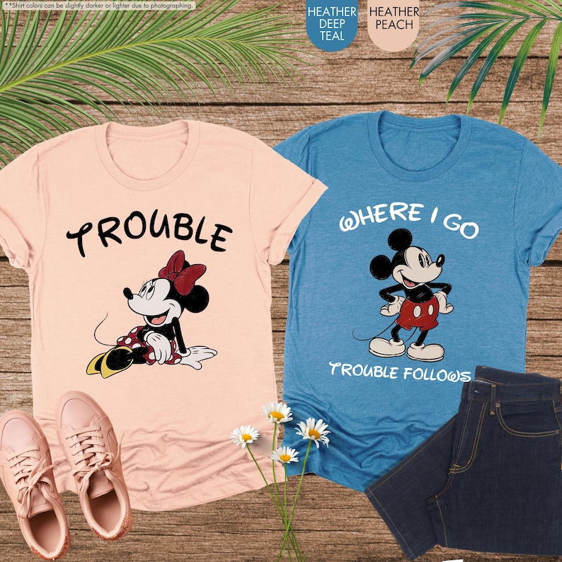 May include: Two t-shirts, one peach and one teal, with Disney character graphics. The peach shirt features Minnie Mouse and the word "TROUBLE." The teal shirt shows Mickey Mouse and the text "WHERE I GO TROUBLE FOLLOWS." A pair of pink sneakers and jeans are also visible.