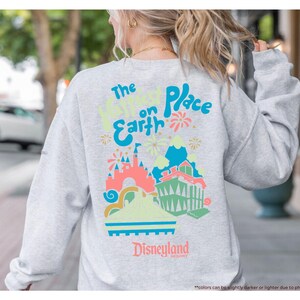 Pu&ograve; includere: Una felpa grigia con una stampa grafica colorata che dice "The Happiest Place on Earth" e presenta un'immagine di cartone animato del castello di Cenerentola e fuochi d'artificio. Il testo "Disneyland" &egrave; stampato sotto la grafica.