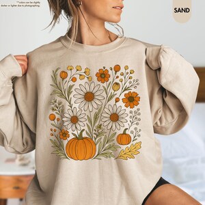 May include: A sand-coloured sweatshirt with a floral design. The design features daisies, orange flowers, and pumpkins, with green leaves and stems. The word "SAND" is printed in a small, rounded font in the upper right corner.
