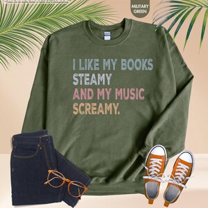 May include: Military green sweatshirt with the text "I LIKE MY BOOKS STEAMY AND MY MUSIC SCREAMY." The image also shows a pair of dark wash jeans, orange sneakers, and a pair of eyeglasses.