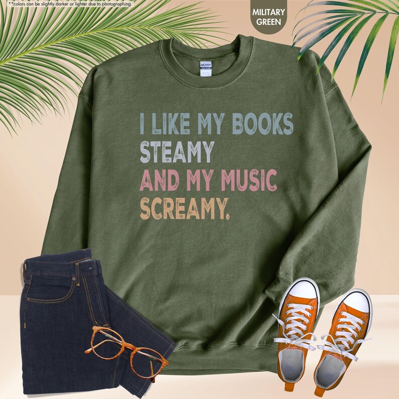 May include: Military green sweatshirt with the text "I LIKE MY BOOKS STEAMY AND MY MUSIC SCREAMY." The image also shows a pair of dark wash jeans, orange sneakers, and a pair of eyeglasses.