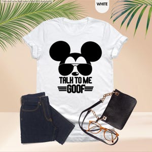 May include: White t-shirt with a black Mickey Mouse graphic wearing sunglasses and the text "TALK TO ME GOOF". The image includes dark blue jeans, a black purse, and a pair of eyeglasses.