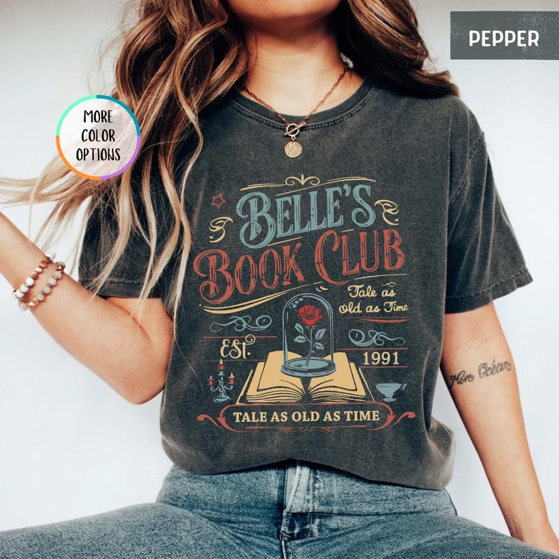 May include: A dark gray t-shirt with a vintage design. The shirt features the text "Belle's Book Club" in blue and red, along with the phrase "Tale as old as Time" and the year "1991". A rose in a glass dome is also depicted.