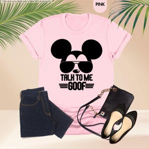 May include: A pink t-shirt featuring a black Mickey Mouse graphic with sunglasses and the text "TALK TO ME GOOF". The image also shows dark blue jeans, a black handbag, and black flat shoes.