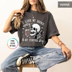 May include: Dark grey t-shirt featuring a skull graphic reading a book and holding a coffee cup. The text says "I like my books spicy & my coffee icy". The t-shirt has a relaxed fit.