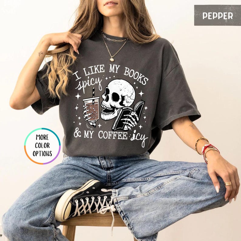 May include: Dark grey t-shirt featuring a skull graphic reading a book and holding a coffee cup. The text says "I like my books spicy & my coffee icy". The t-shirt has a relaxed fit.