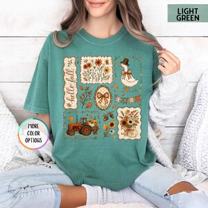 May include: A light green t-shirt featuring a graphic print with the text "hello fall" and various autumn-themed illustrations including a tractor, a duck wearing a hat, sunflowers, pumpkins, acorns, a plaid bow, and a festive bunting banner.