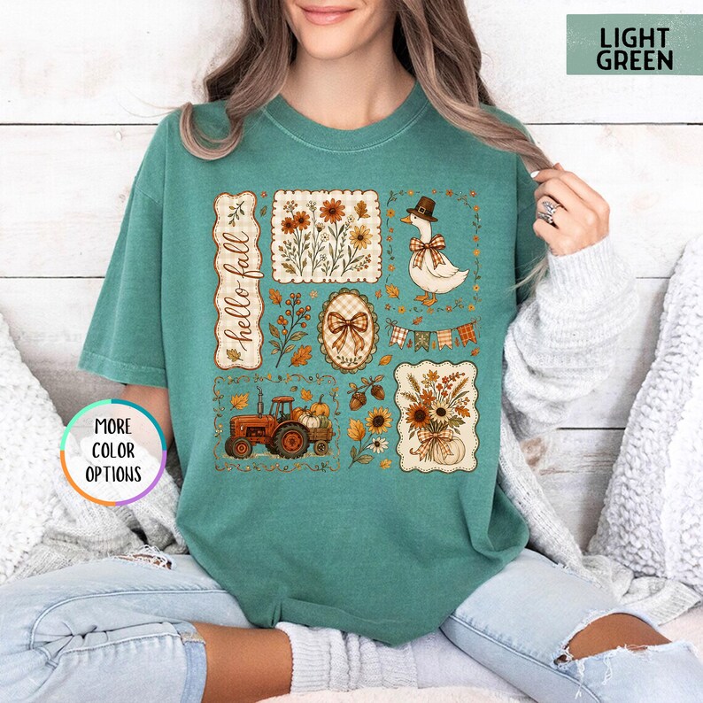 May include: A light green t-shirt featuring a graphic print with the text "hello fall" and various autumn-themed illustrations including a tractor, a duck wearing a hat, sunflowers, pumpkins, acorns, a plaid bow, and a festive bunting banner.