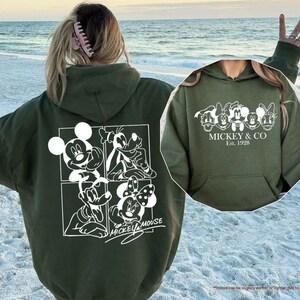 May include: Military green hoodie featuring a white graphic of Mickey Mouse, Goofy, Minnie Mouse, and Donald Duck. The back displays a four-panel design with Mickey Mouse and Minnie Mouse. The front has the text "MICKEY & CO Est. 1928".