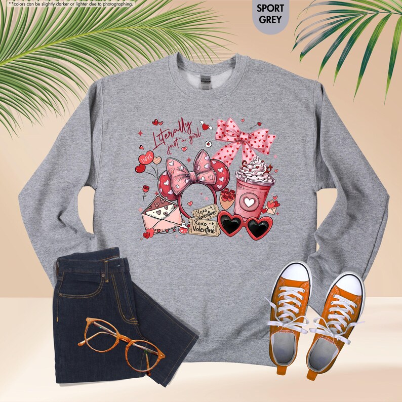 May include: A sport grey sweatshirt featuring a Valentine's Day design with a pink bow, heart-shaped sunglasses, and a coffee cup. The text reads "Literally just a girl." The image also shows dark blue jeans, orange trainers, and glasses.