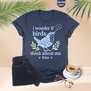 May include: A heather navy blue short-sleeve t-shirt featuring a light blue graphic of a bird and the text "i wonder if birds think about me too" with a small heart in a thought bubble, accompanied by yellow leaf accents and four-pointed stars.