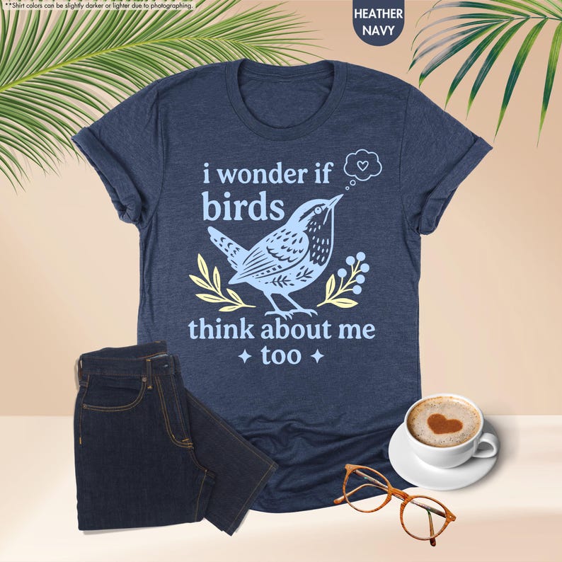 May include: A heather navy blue short-sleeve t-shirt featuring a light blue graphic of a bird and the text "i wonder if birds think about me too" with a small heart in a thought bubble, accompanied by yellow leaf accents and four-pointed stars.
