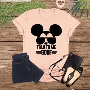 May include: A heather peach t-shirt featuring a black Mickey Mouse graphic wearing sunglasses and the text "TALK TO ME GOOF." The image also shows dark blue jeans, a black handbag, and black flats.