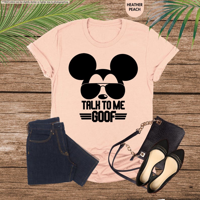 May include: A heather peach t-shirt featuring a black Mickey Mouse graphic wearing sunglasses and the text "TALK TO ME GOOF." The image also shows dark blue jeans, a black handbag, and black flats.
