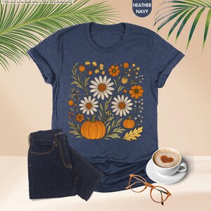 May include: Navy blue heather t-shirt with a floral design including white daisies, orange pumpkins, and autumn elements. Dark blue jeans are next to the shirt. A coffee cup and glasses are also visible.