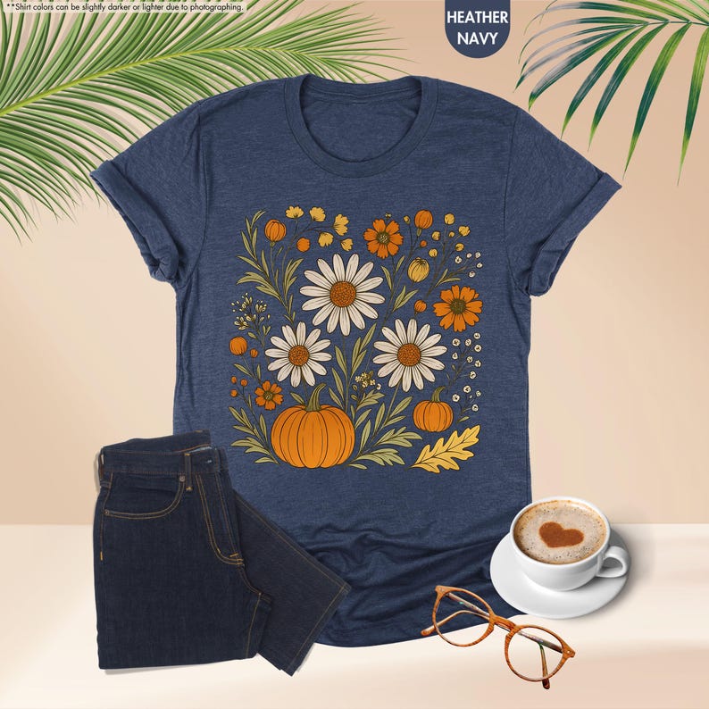 May include: Navy blue heather t-shirt with a floral design including white daisies, orange pumpkins, and autumn elements. Dark blue jeans are next to the shirt. A coffee cup and glasses are also visible.