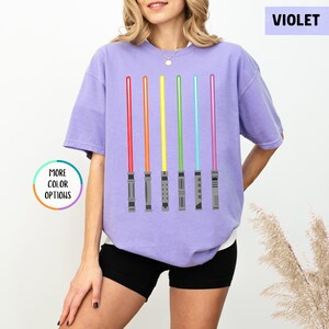 K&ouml;nnte beinhalten: Violettes T-Shirt mit einer Grafik von sechs farbigen Lichtschwertern in Rot, Orange, Gelb, Gr&uuml;n, Blau und Lila. Das Shirt hat den Text "MORE COLOR OPTIONS" in einem Kreis und "VIOLET" in einem Rechteck.