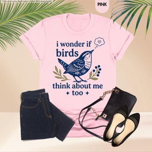 May include: A light pink short-sleeved t-shirt featuring a dark blue graphic of a bird with a thought bubble containing a heart. The text reads, "i wonder if birds think about me too." The shirt is paired with dark blue denim jeans and a black purse.